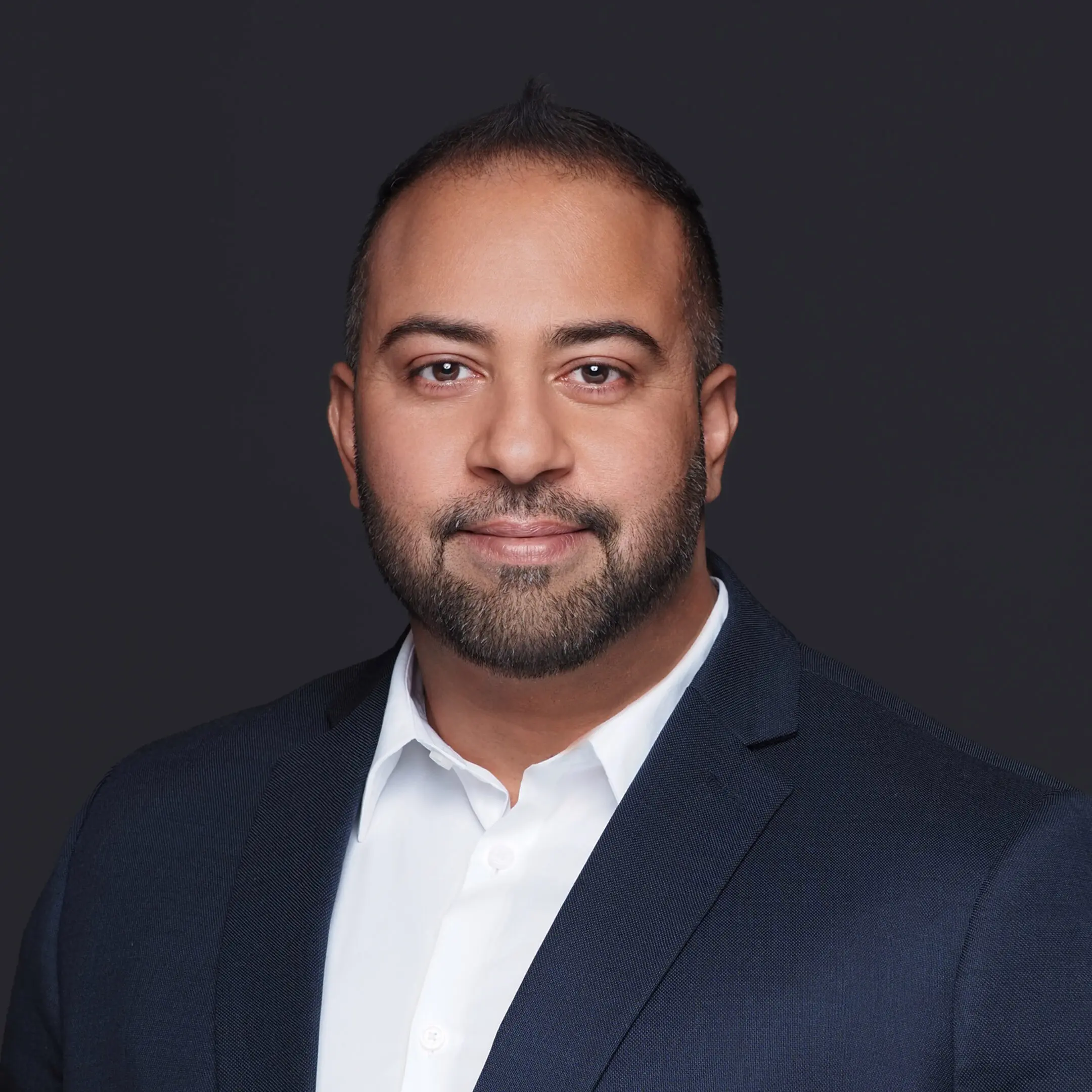 67d1f9a77a0e6_Robin Brar square crop A man in a suit and tie standing up against a black background.